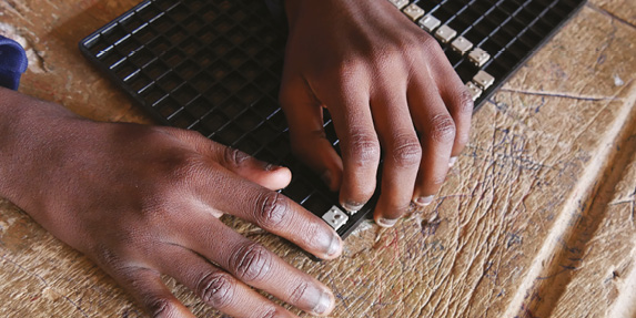 Two hands of a Black child placed on a worn wooden table. His hands are composing a word in Braille on a plastic frame